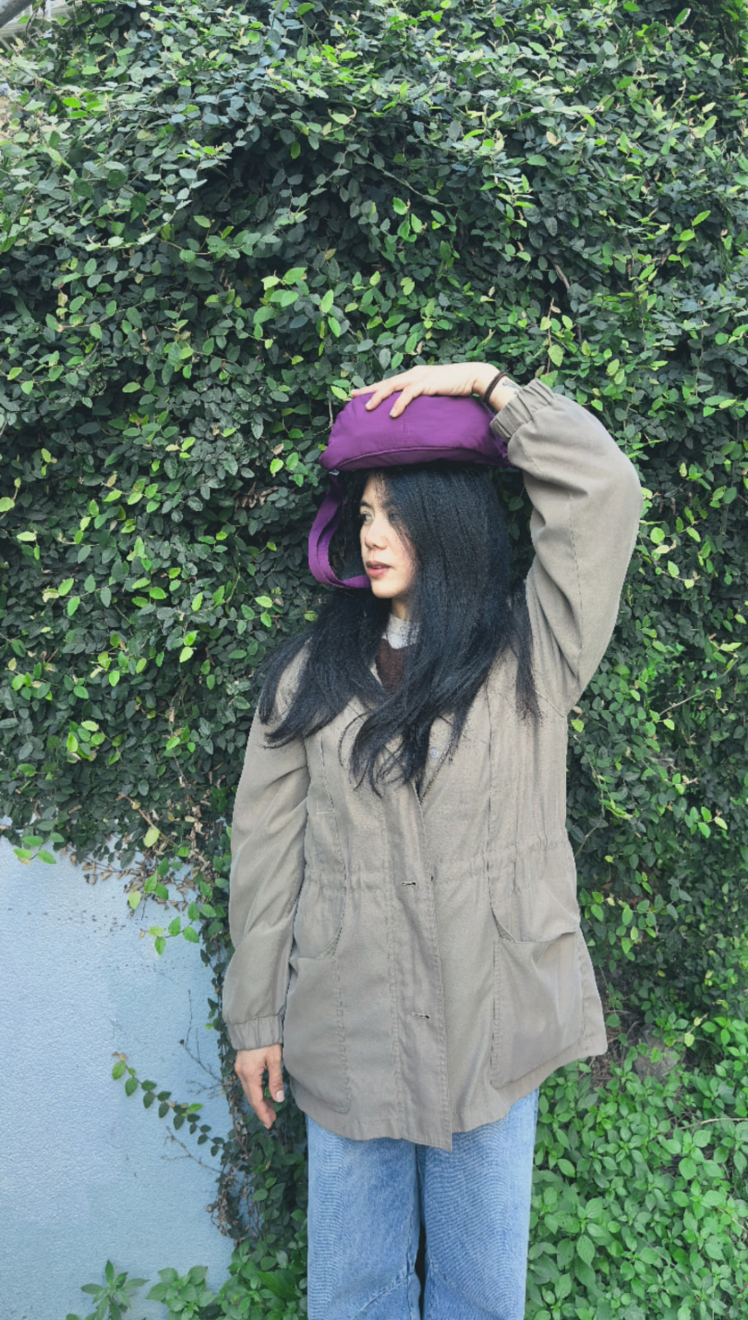 Let standing in front of leafy greenery, balancing a purple bag on her head